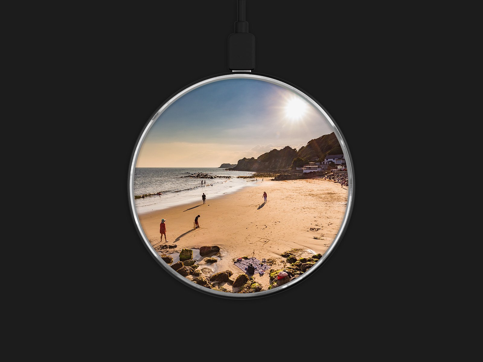 A wireless charging pad displays a beach scene, with people walking on sand near the shoreline, rocky cliffs in the background, and the sun shining in a clear sky. The pad is set against a black background.